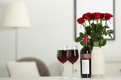 Bottle, glasses of red wine and vase with roses on white table in room, space for text. Romantic date