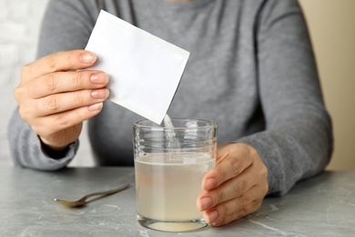 Woman pouring powder from medicine sachet into glass of water on grey marble table, closeup