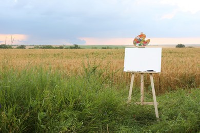Wooden easel with blank canvas and painting equipment in field. Space for text