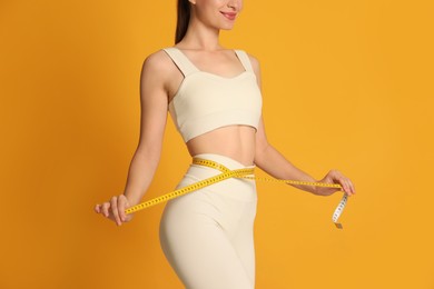 Happy woman in sportswear measuring waist with tape on yellow background, closeup