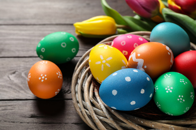 Colorful Easter eggs in decorative nest and tulips on wooden background, closeup