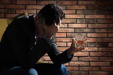 Addicted man with glass of alcoholic drink in armchair near red brick wall
