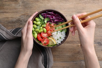 Woman eating poke bowl with salmon, edamame beans and vegetables at wooden table, top view