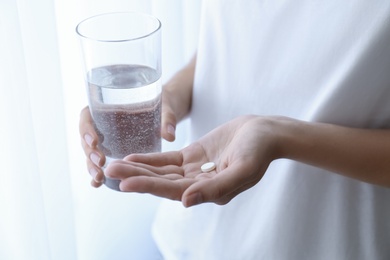 Woman holding glass of water and pill near window at home, closeup