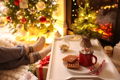 Photo of Woman sitting near fireplace at home, focus on tray with cocoa and snacks. Cozy winter holidays atmosphere