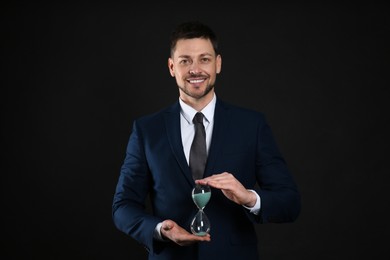 Happy businessman holding hourglass on black background. Time management