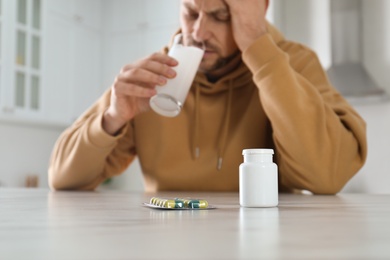 Man taking medicine for hangover at table in kitchen, focus on bottle with pills