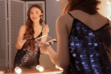 Photo of Beautiful young woman in elegant dress applying blusher with brush near mirror indoors