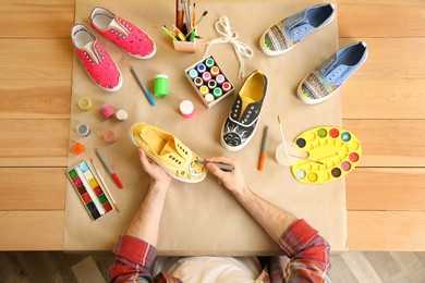 Man painting on sneaker at wooden table, top view. Customized shoes