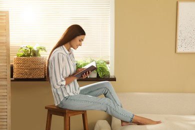 Beautiful young woman reading book on stool at home