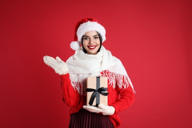 Woman in Santa hat, knitted mittens, scarf and sweater holding Christmas gift on red background