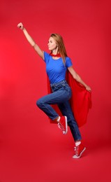 Confident woman in superhero cape jumping on red background