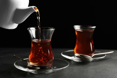Pouring traditional Turkish tea from pot into glass on black table, closeup