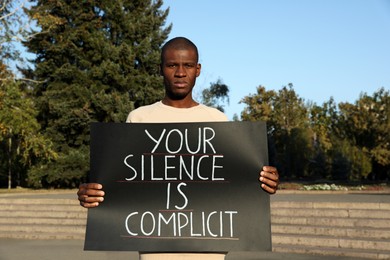 Photo of African American man holding sign with phrase Your Silence Is Complicit outdoors. Racism concept