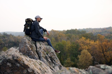Hiker with travel backpack sitting on steep cliff on autumn day