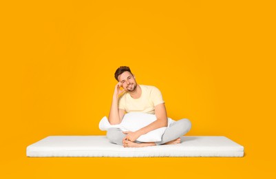Smiling man with pillow sitting on soft mattress against orange background