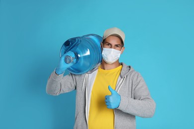 Courier in medical mask with bottle for water cooler showing thumb up on light blue background. Delivery during coronavirus quarantine