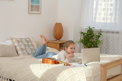 Photo of Little girl learning to play ukulele with online music course at home. Time for hobby