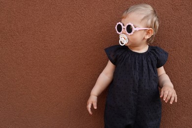 Cute little girl wearing stylish clothes with sunglasses and pacifier near brown wall. Space for text