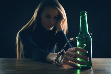 Alcohol addiction. Woman handcuffed to bottle of beer at wooden table against black background, focus on hand