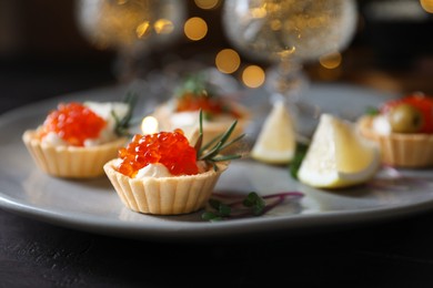 Photo of Delicious tartlets with red caviar, cream cheese and lemon served near wine on black table, closeup