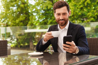 Handsome bearded man with cup of drink and smartphone at table in cafe