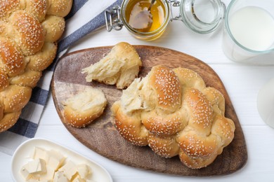 Photo of Homemade braided bread with sesame seeds, butter, milk and honey on white wooden table, flat lay. Traditional challah
