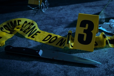 Yellow tape, crime scene marker and bloody knife on grey stone table at night