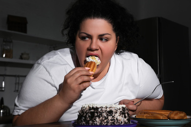 Depressed overweight woman eating sweets in kitchen at night