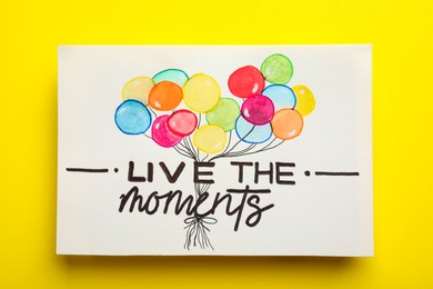 Card with life-affirming phrase Live The Moments on yellow background, top view