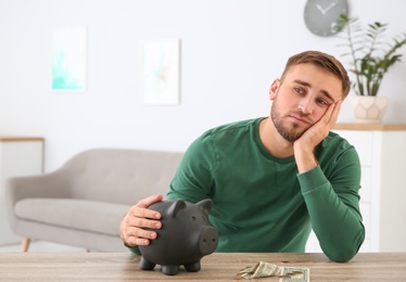 Young man with piggy bank and money at table indoors. Space for text
