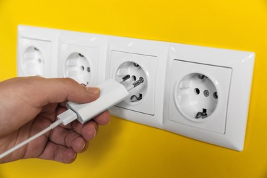 Woman plugging charger into power socket on yellow wall, closeup. Electrical supply