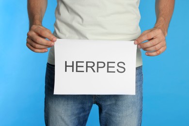 Photo of Man holding paper with word Herpes on light blue background, closeup
