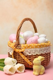 Photo of Wicker basket with festively decorated Easter eggs, bunny and beautiful tulips on pink table against color textured background