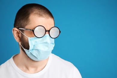 Man with foggy glasses caused by wearing disposable mask on blue background, space for text. Protective measure during coronavirus pandemic