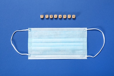 Photo of Medical face mask and word Distance made with small wooden cubes on blue background, flat lay