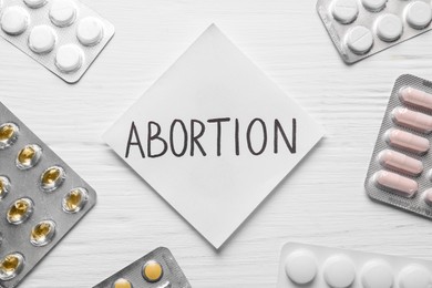 Paper note with word Abortion and pills on white wooden background, flat lay