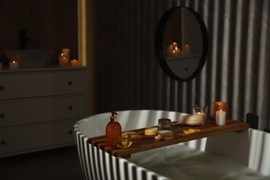 Wooden bath tray with candles and bathroom amenities on tub indoors