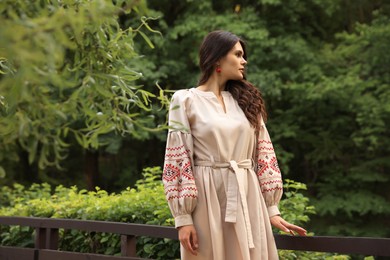 Beautiful woman wearing embroidered dress near wooden railing in countryside. Ukrainian national clothes