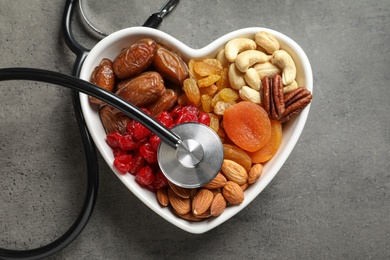 Heart shaped bowl with dried fruits, nuts and stethoscope on grey background, top view
