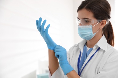 Doctor in protective mask and glasses putting on medical gloves against light background