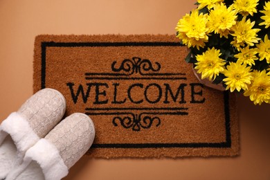 Photo of Doormat with word Welcome, slippers and flowers on brown background, flat lay