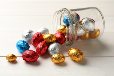 Overturned glass jar with chocolate eggs wrapped in colorful foil on white wooden table