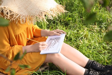 Photo of Young woman drawing with pencil in notepad while sitting on green grass