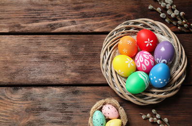Colorful Easter eggs in decorative nest and willow branches on wooden background, flat lay. Space for text