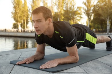 Sporty man doing plank exercise on mat near river