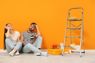 Photo of Tired designers sitting on floor with painting equipment near freshly painted orange wall indoors