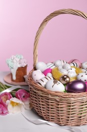 Photo of Wicker basket with festively decorated Easter eggs and beautiful tulips on white marble table against pink background
