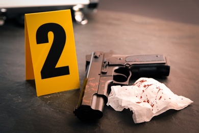 Napkin with blood, gun and evidence marker on black slate table, closeup. Crime scene