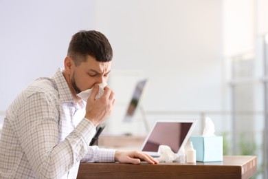Photo of Ill man at table with nasal spray, drops and box of paper tissues in office